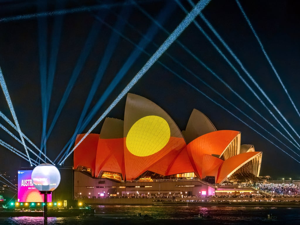 The Sydney Opera House is illuminated at night with vibrant red and yellow Indigenous flag art projections. Blue laser beams radiate into the night sky.