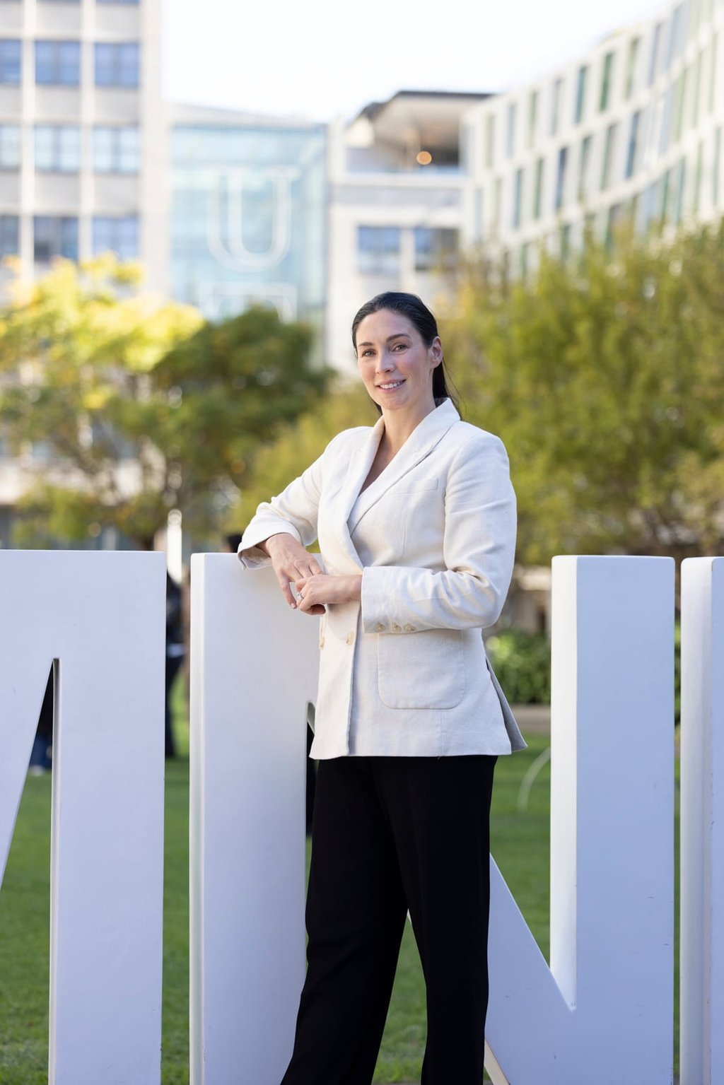 Samantha, wearing a white blazer, stands in Alumni Green, leaning against the letter N.