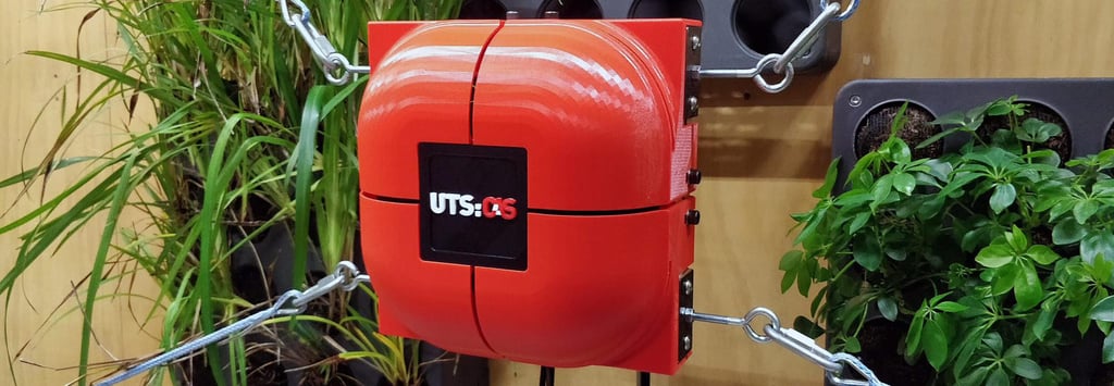 Red robotic device with "UTS:RS" logo, mounted on a wooden wall. Surrounded by green plants, connected with metal cables, conveying a tech-nature blend.