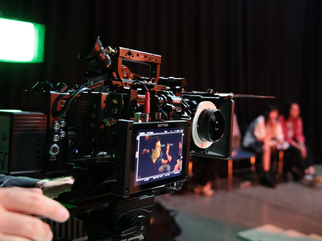 Close-up of a professional video camera on a film set, focused on two people in a dimly lit room. The atmosphere is creative and focused.