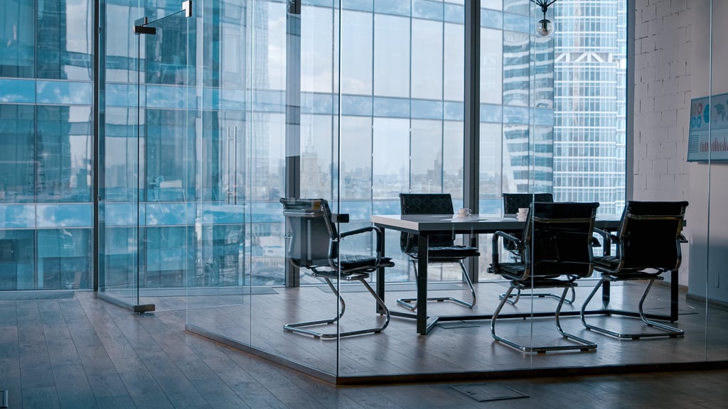 Empty meeting room in office, six black leather chairs are placed around a glass table while skyscrapers can be viewed in the background.