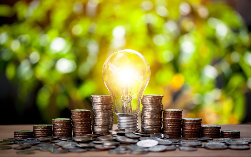 A glowing light bulb surrounded by stacks of coins on a table, set against a blurred green and yellow background.