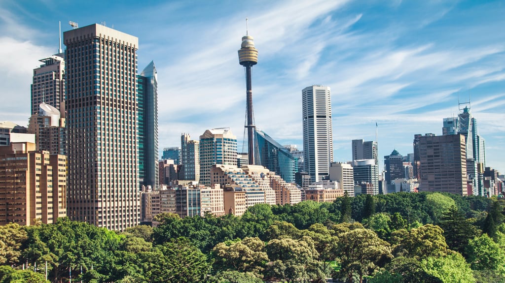 Skyline of Sydney featuring tall modern skyscrapers and a distinctive tower under a clear blue sky. Lush green park in the foreground, conveying vibrancy.