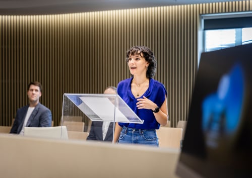 A woman stands at a podium, presenting to an audience seated in rows, engaged and attentive.