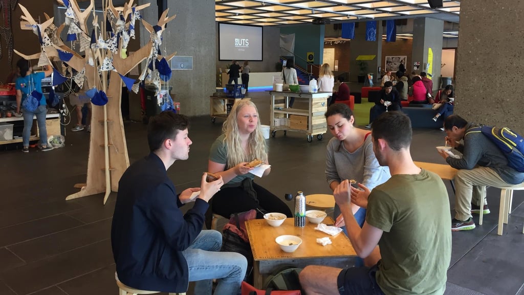 A group of four people sits around a small wooden table, eating and chatting in a modern, busy communal space. A decorative paper tree stands nearby.