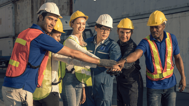 A diverse group of six construction workers in safety gear stand united in a circle with hands stacked, conveying teamwork and solidarity.