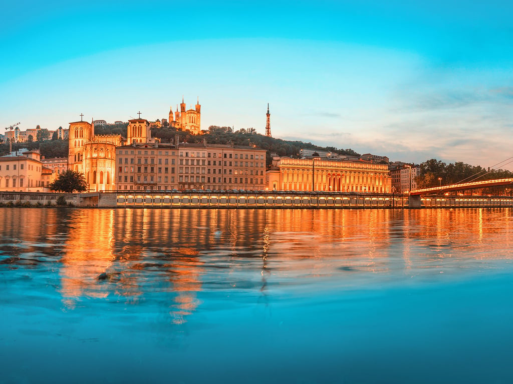 Illuminated cityscape of Lyon at dusk, featuring historic buildings on the waterfront and a modern suspension bridge, reflected in the calm river.