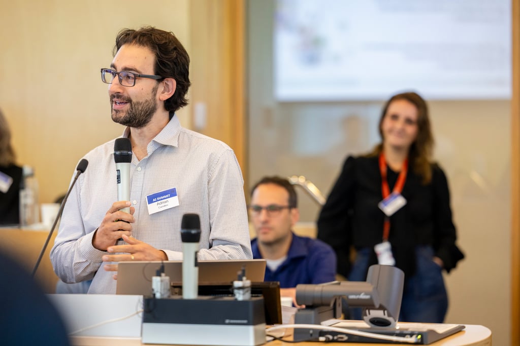 Dr Adrian Camilleri speaks into a handheld microphone at the Marketing in AI Summit, standing behind a lectern with presentation equipment, while other attendees listen in the background.