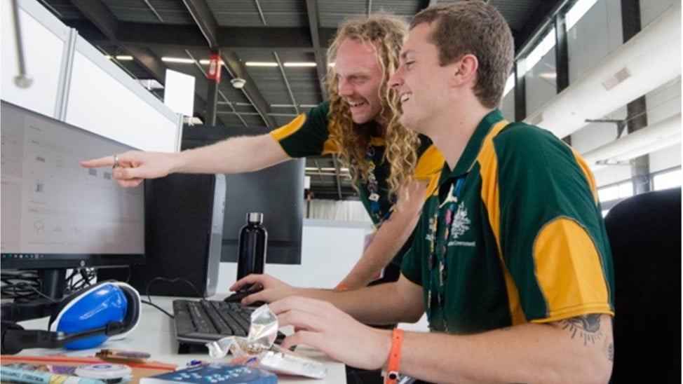 Two people in matching green and yellow shirts smiling at a computer screen. One points at the monitor, suggesting a collaborative and positive atmosphere.