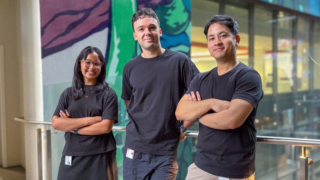 Three people in black shirts stand with arms crossed, smiling, in front of a vibrant mural. The atmosphere is casual and friendly.