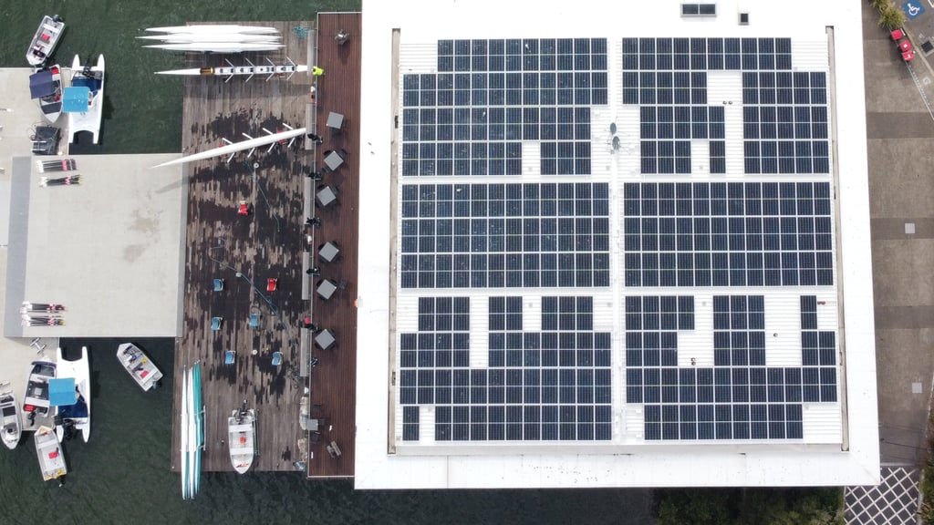 Aerial view of a waterside building with a solar panel roof. Adjacent dock holds boats, kayaks, and outdoor seating. Nearby road has moving cars.