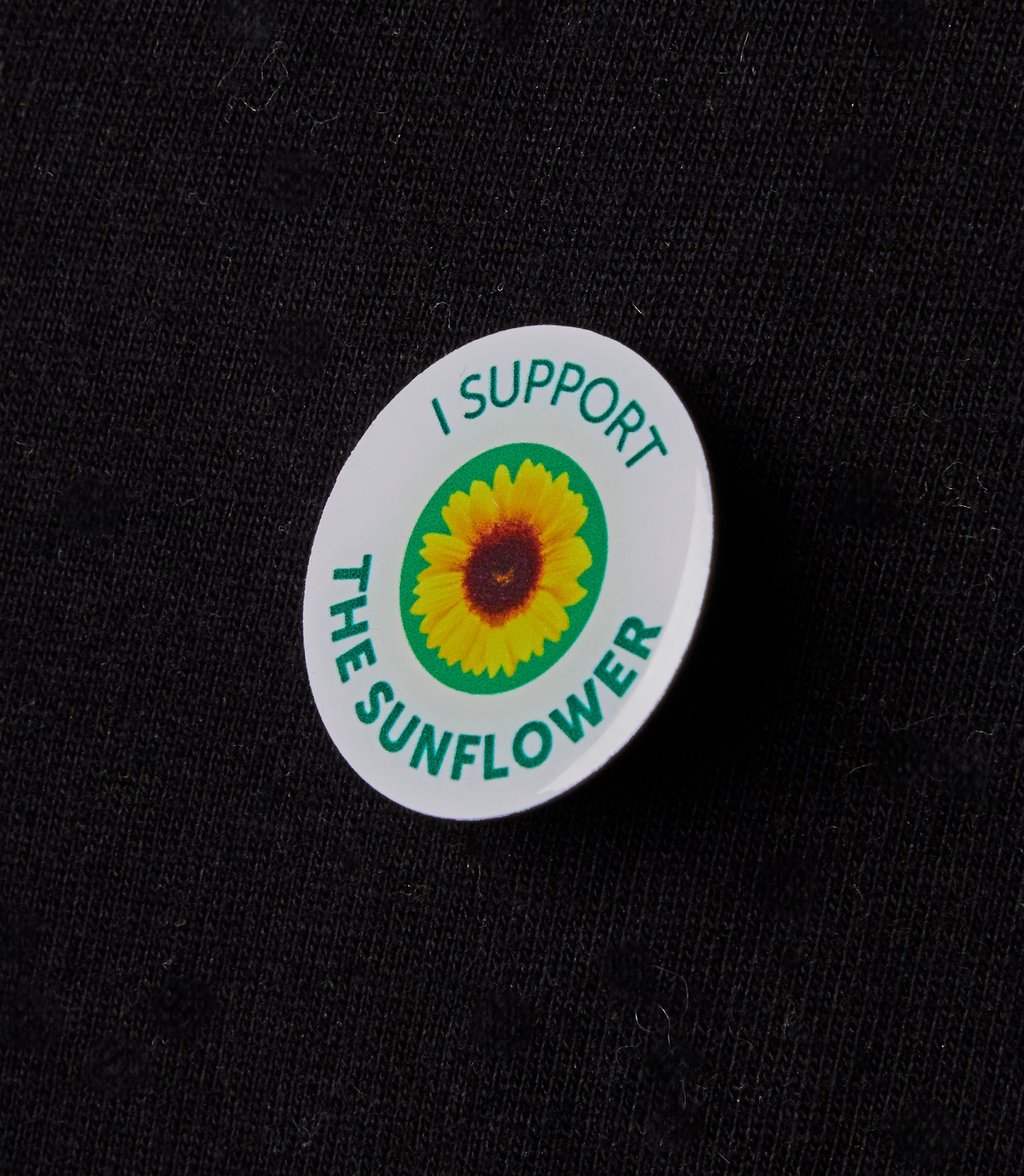A round badge on a black fabric surface features a sunflower illustration with the text "I Support the Sunflower" encircling it in green letters.