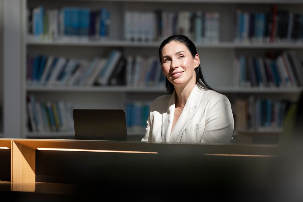 Samantha, wearing a white blazer, sits in the library with a bright beam of sunlight shining on her.