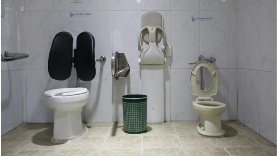 Public restroom with white tiled walls features an adult toilet with black backrests, a child-sized toilet, a wall-mounted baby seat, and a green trash can.