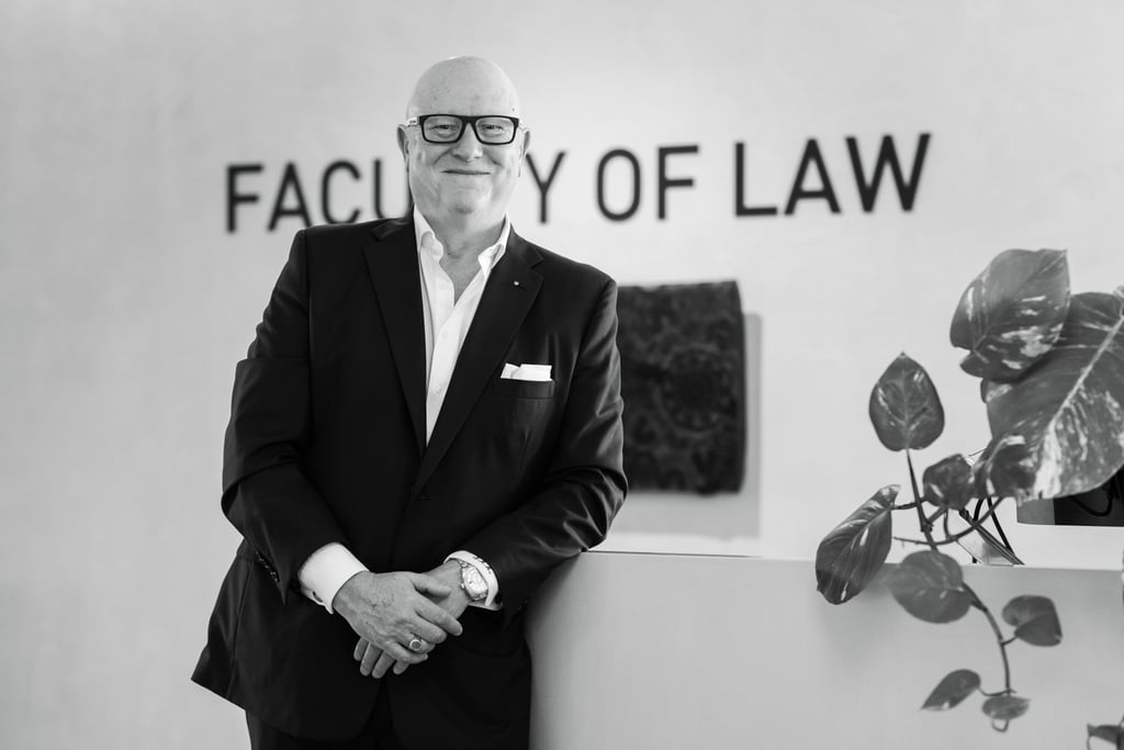 Black and white photo of Nicholas. He is wearing a dark-coloured suit and leaning on a desk in front of a Faculty of Law sign.