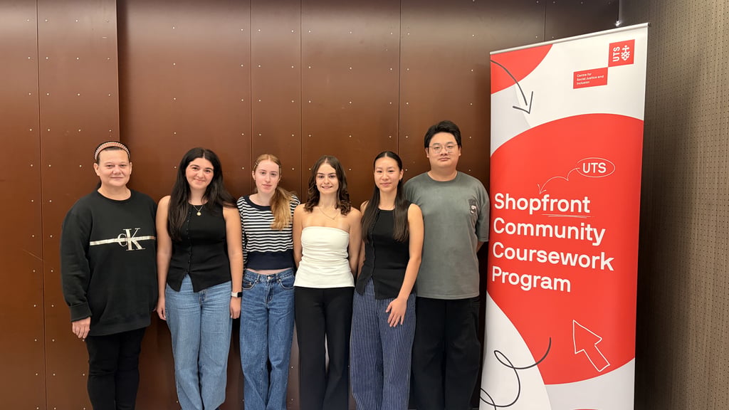 Six students standing in a row next to a red banner reading ‘UTS Shopfront Community Coursework Program’ against a brown wall.