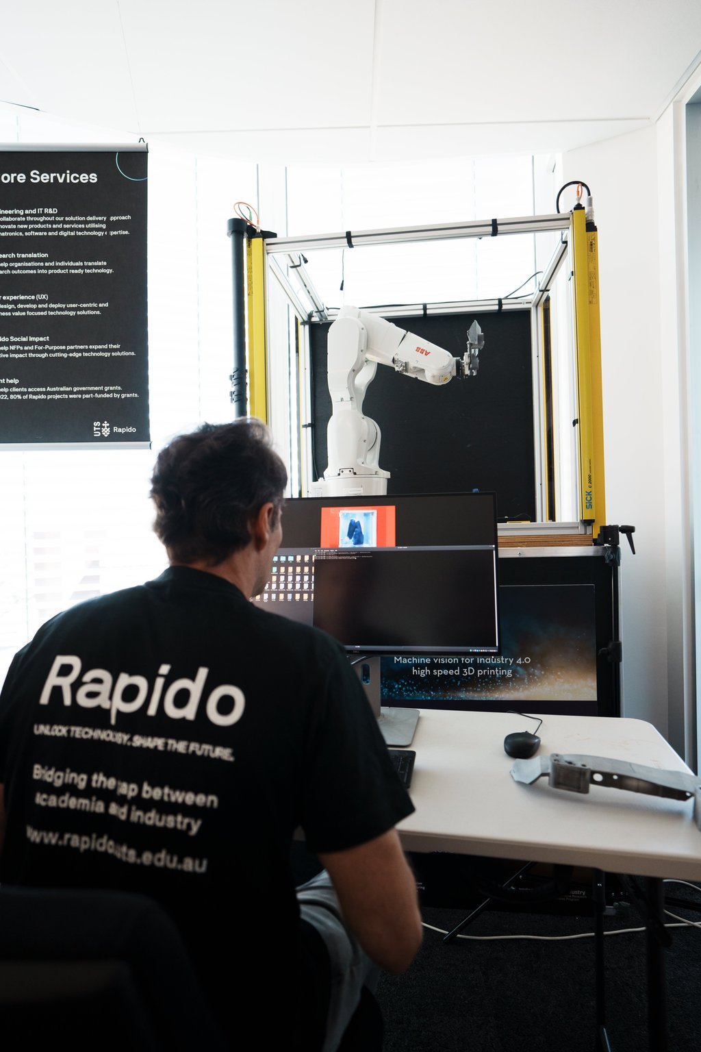 UTS Rapido team member operating a robotic arm for machine vision and 3D printing research, with a services banner in the background