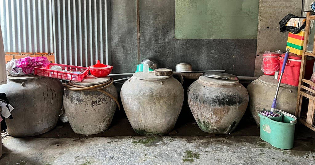 Five large, aged clay pots with lids are lined against a corrugated metal and green wall. Nearby, red and pink items, and a broom sit on a shelf.