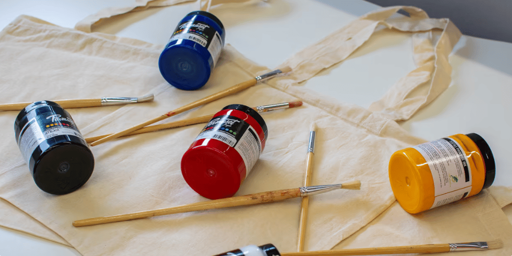 Fabric tote bags with paint pots and brushes arranged on top for a painting activity.