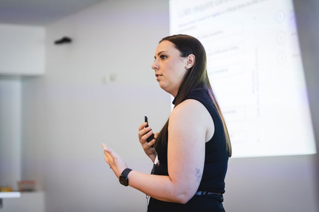 In the foreground, a female speaker in a black dress is gesticulating in one hand and holding a buzzer in the other as she addresses an audience we cannot see. Behind her in the background is a PowerPoint presentation whose slides projected onto the wall.