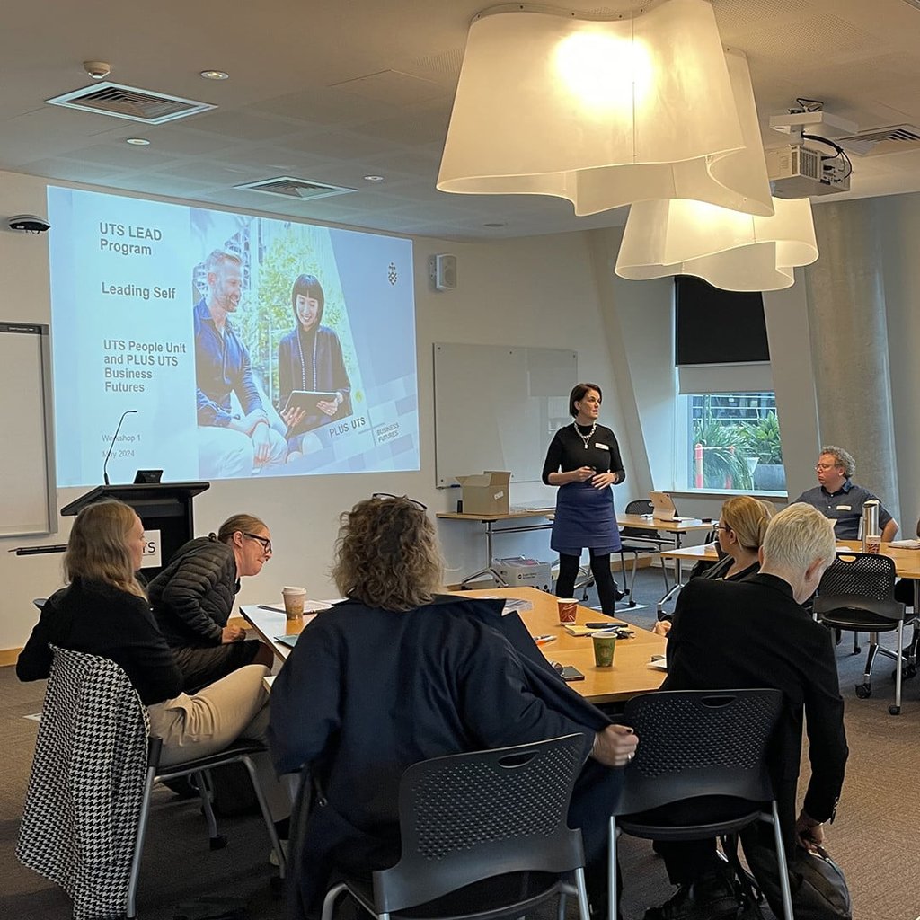 A group of people in a conference room attends a presentation. A woman stands at the front, speaking next to a screen. Participants sit around tables, engaging and taking notes. The atmosphere is focused and collaborative.