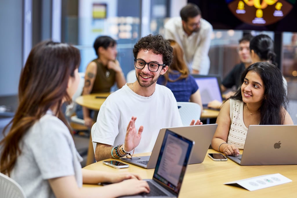 A diverse group of people collaborate at a round table with laptops. A smiling man gestures while two women listen, creating a lively, interactive atmosphere.