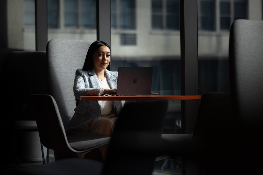 Arhita, wearing a light blue blazer, sits on a grey seat working at her laptop
