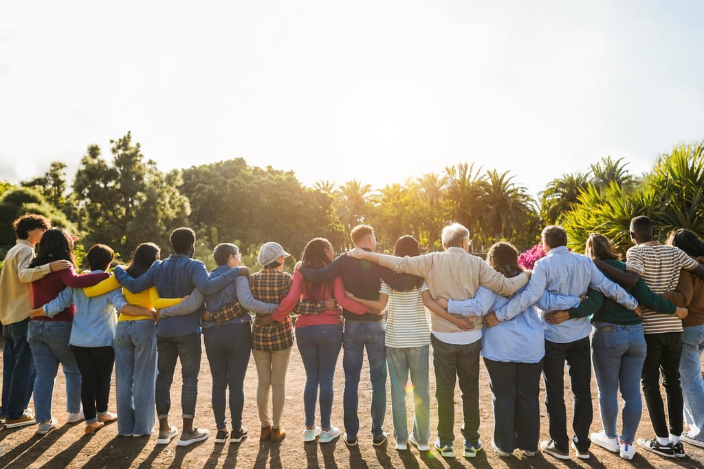 A diverse group of people stands in a line outdoors, arms around each other, facing the sunset.