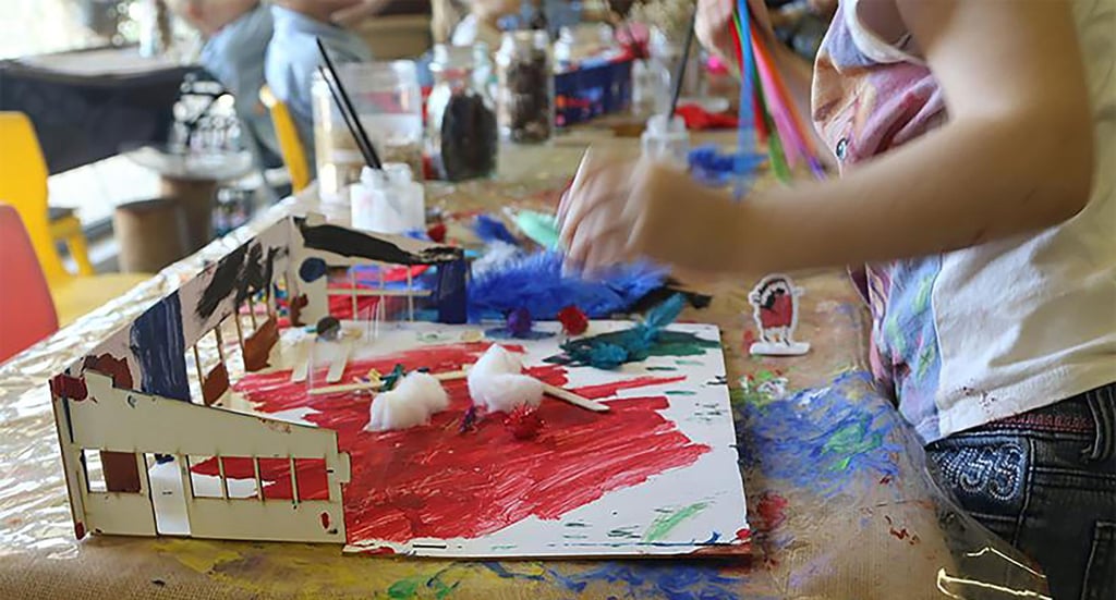Child crafting with colorful feathers, cotton, and red paint on paper. A playful atmosphere with art supplies and vibrant creativity on a wooden table.