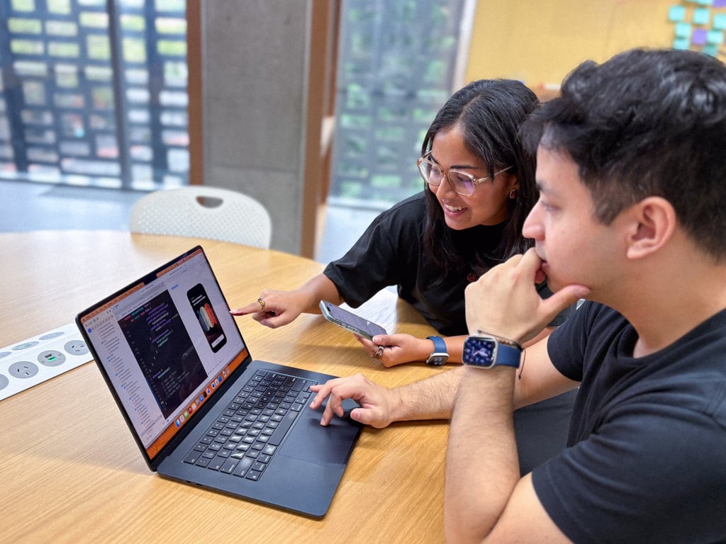 Two people collaborate at a table, reviewing code on a laptop, with an app design open. They appear focused and engaged in discussion.