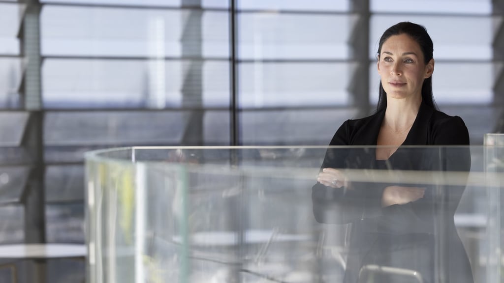 Samantha smiles, looking into the distance, with her arms folded gently. Slatted windows are seen behind her.