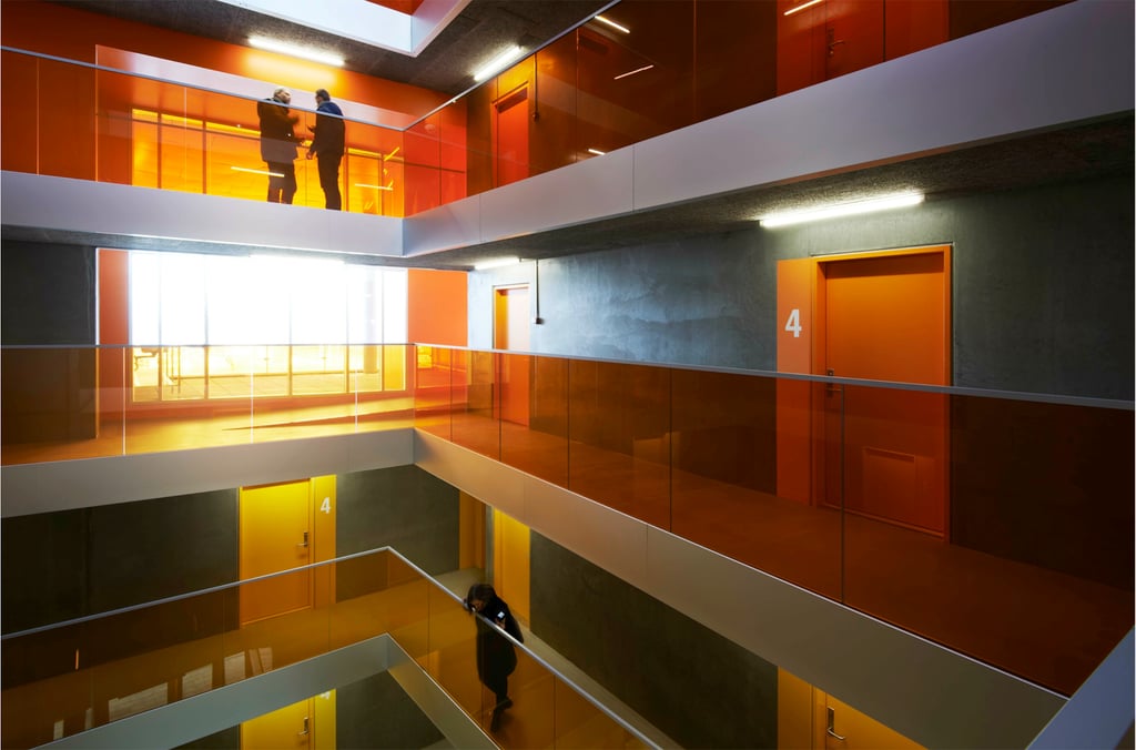 Internal photograph of the Aarhus Student Housing showing several mezzanine floors around the atrium bathed in orange from sunlight coming through the window onto the orange glass panels on the inside of the walkways.