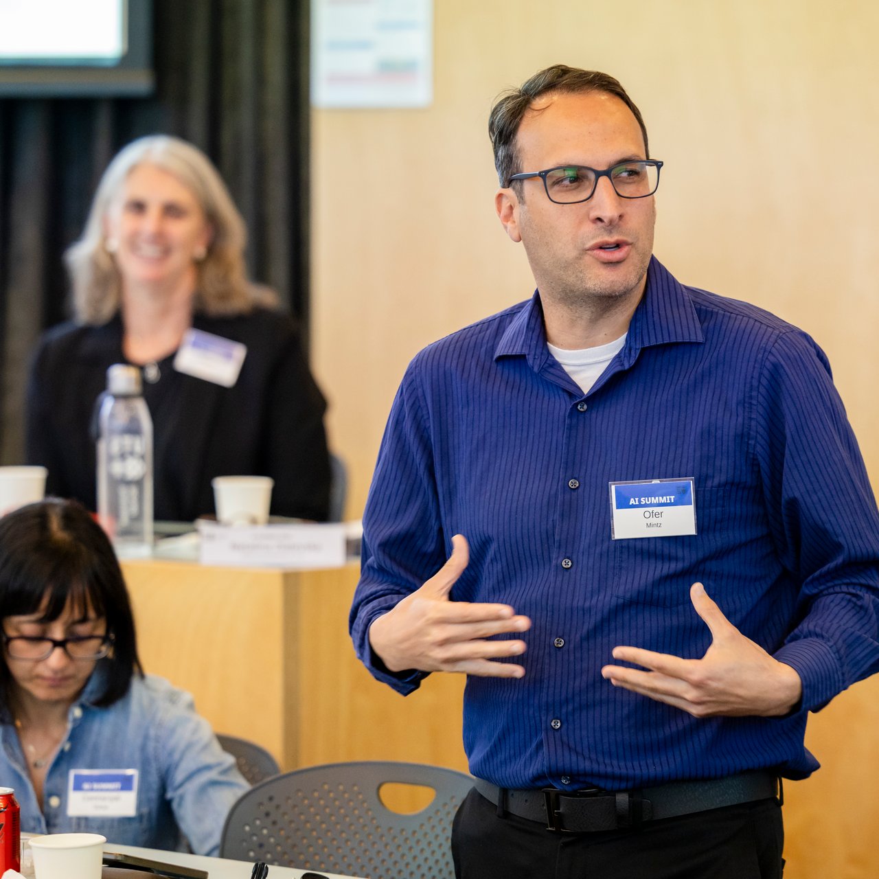 Professor Ofer Mintz is stood in the centre of the oval classroom gesticulating with his hands as he addresses an audience offscreen. He is dressed in a blue top and several attendees can be seen seated behind in listening into the speech.