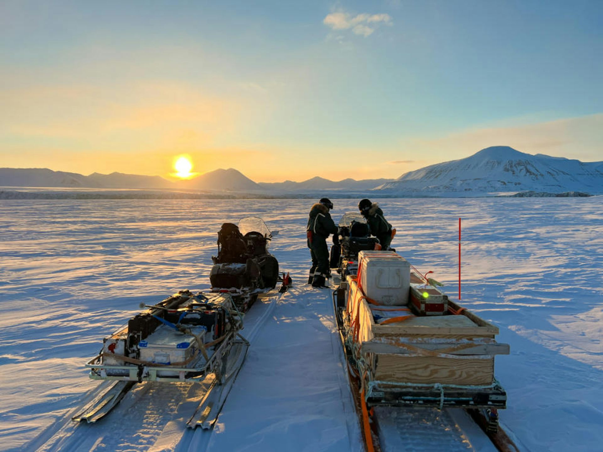 A yellow sunrise over an icy field with people and sleighs laden with scientific equipment