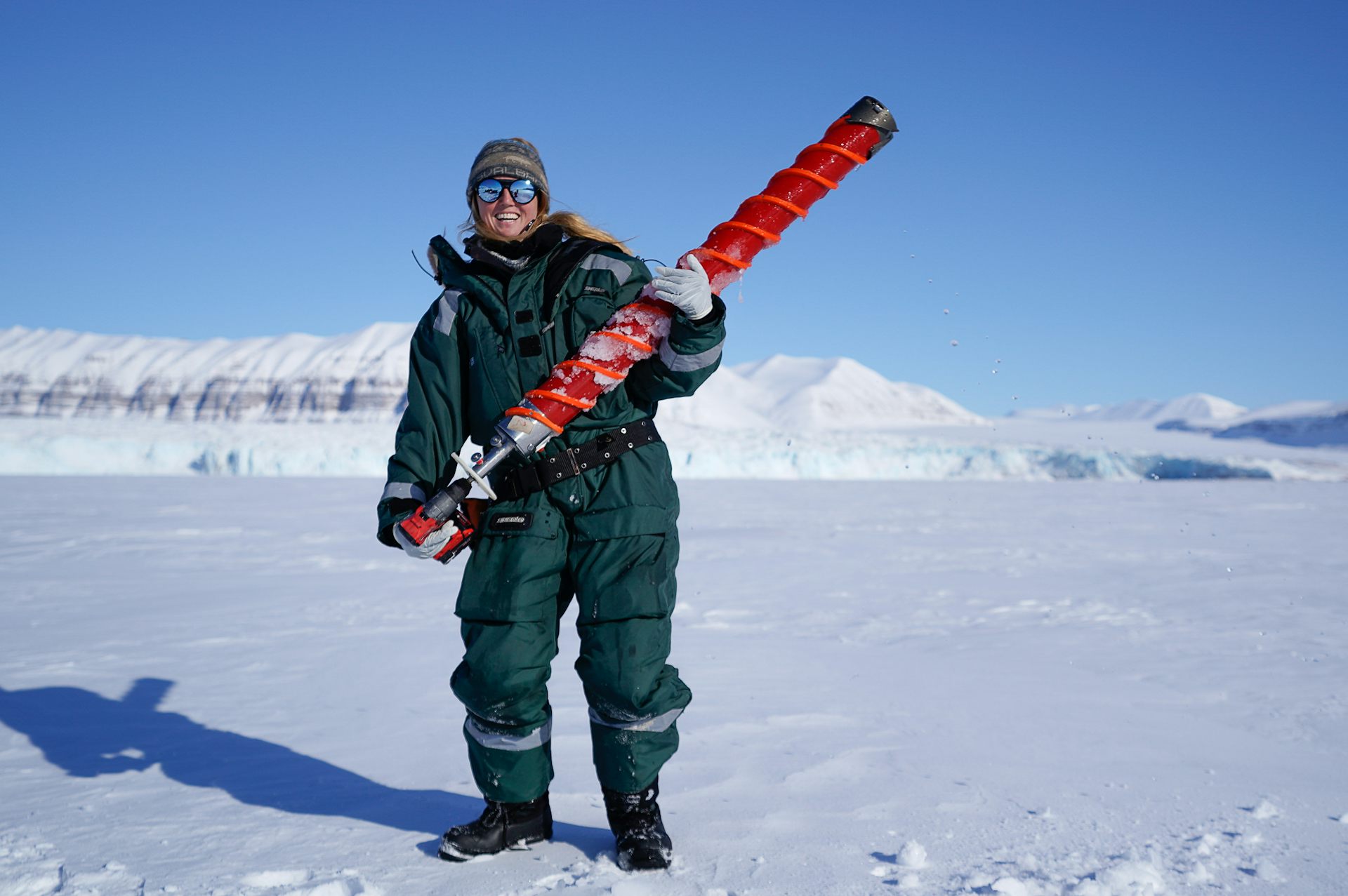 A woman in a green snow suit holds a large red cylinder and smiles at the camera