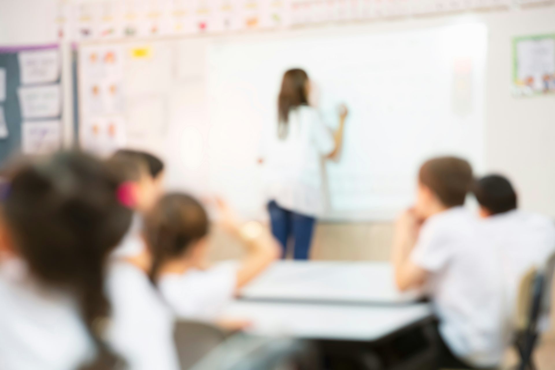 A blurred image of a teacher at a whiteboard with students in white uniform tops seated at desks.