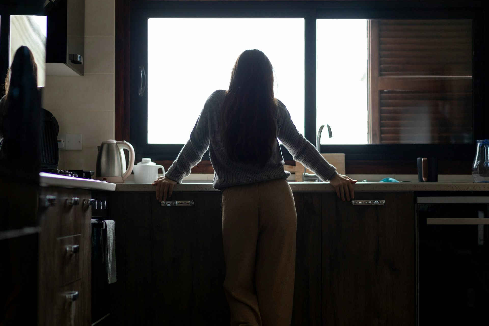 A woman stands alone at her sink, looking out the window.