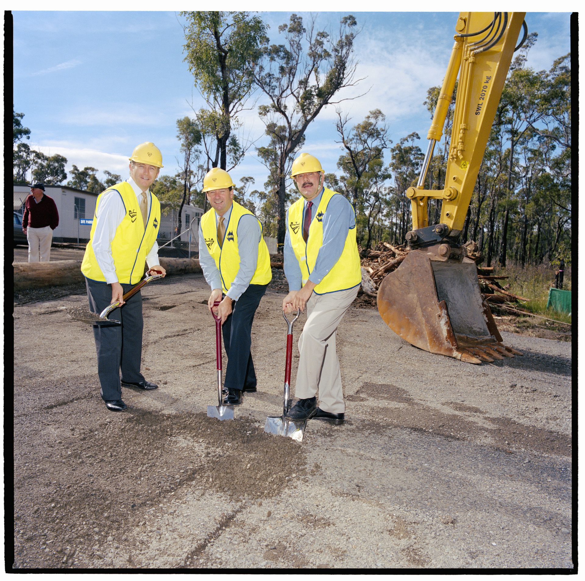 Three politicians wearing business wear and yellow hi-vis vests, with silver shovels, about to dig into dirt at a new construction site