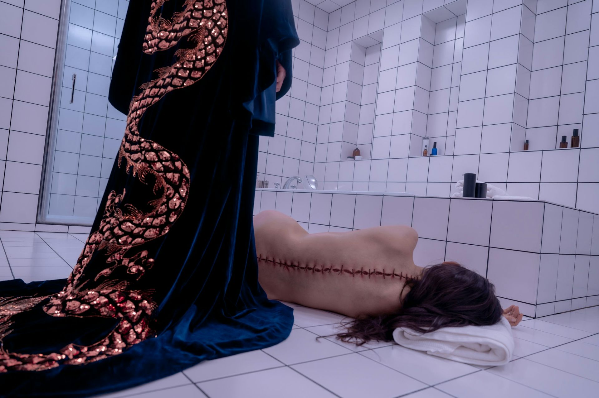 A woman in a dressing gown stands over the body of a woman with a scar down her spine.