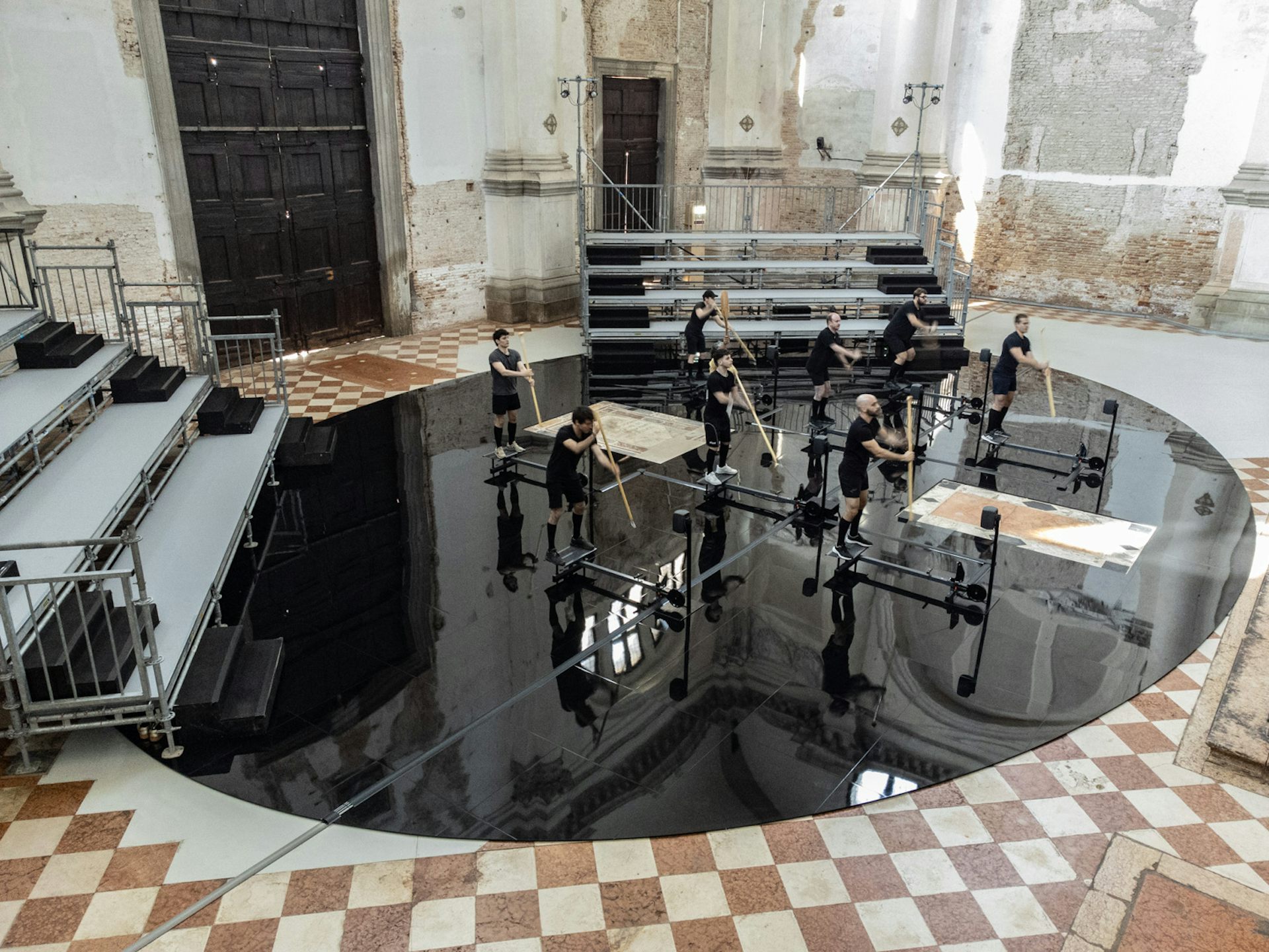 People stand on black boards on a reflective black surface, and paddle the air with oars.