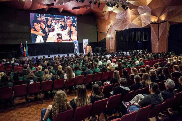 A large amount of high school students sitting in a hall watching a big screen and presenter