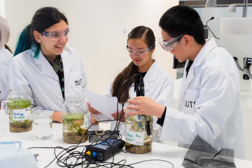 Three high school students standing at a lab table, wearing lab coats, observing an experiment using jars and an electric current