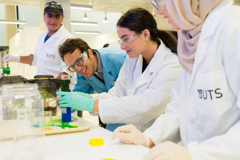 High school students wearing lab coats and safety glasses gather around an experiment