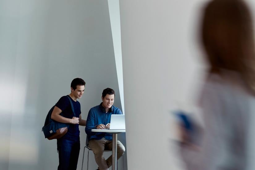 Two people looking at a laptop, one sitting, one standing. In the foreground there is a blurred person walking