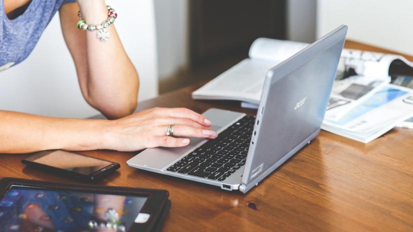 A young woman sits at a table with a laptop, tablet and mobile phone