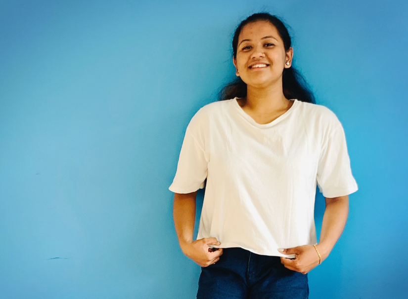 Mathushalini is wearing a white t-shirt and denim shorts, standing against a bright blue background and smiling confidently at the camera