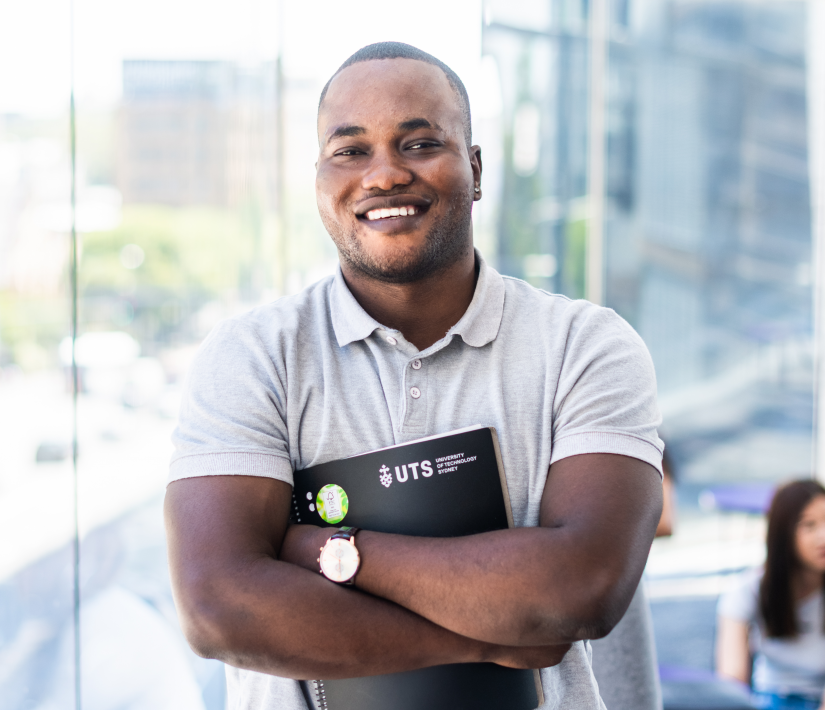 Rock stands, arms folded, with a black folder with 'UTS' written on it against his chest. He wears a white t-shirt. Behind him is a pale background looking out onto the UTS campus through a window.