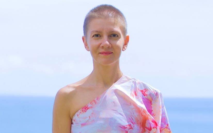 Christina Giarmatzi standing in front of the ocean and wearing a floral off-the-shoulder summer dress and
