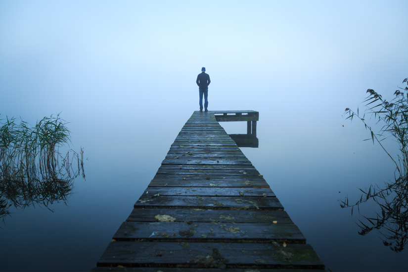A man stands at the end of a jetty. The body of water sits under fog. Some water plants are visible on the left and right of the image.