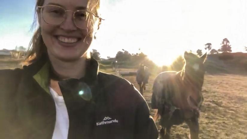 A young woman smiles at the camera, the sun and horses in the background.